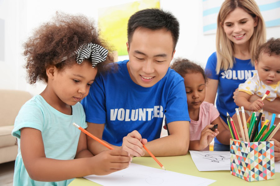 Young man volunteering at a daycare helping a young girl draw.