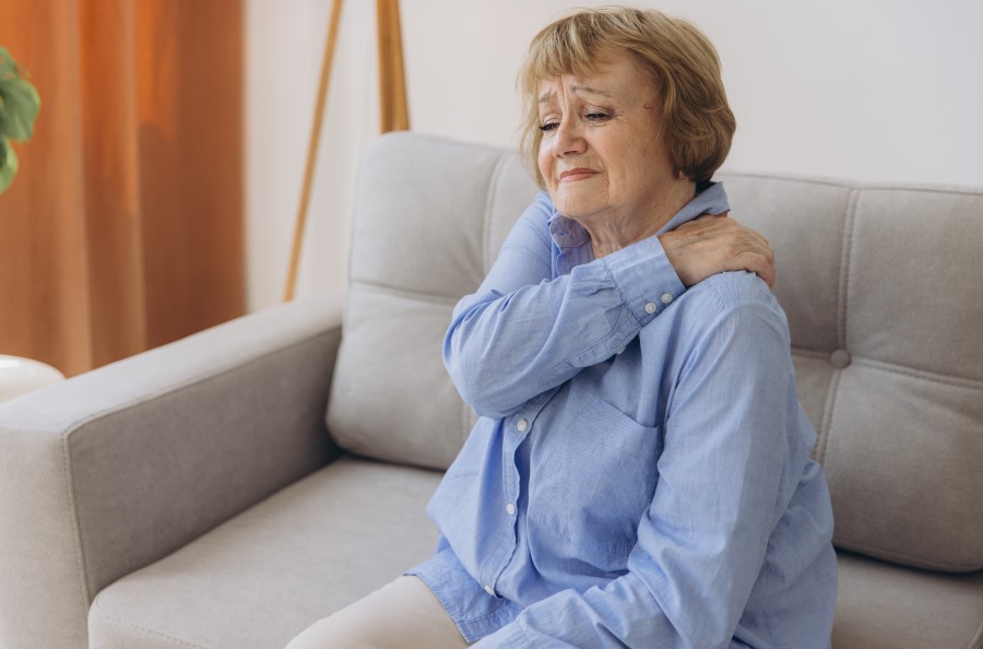Woman sitting on a couch holding left shoulder wondering if her pain could mean a heart attack