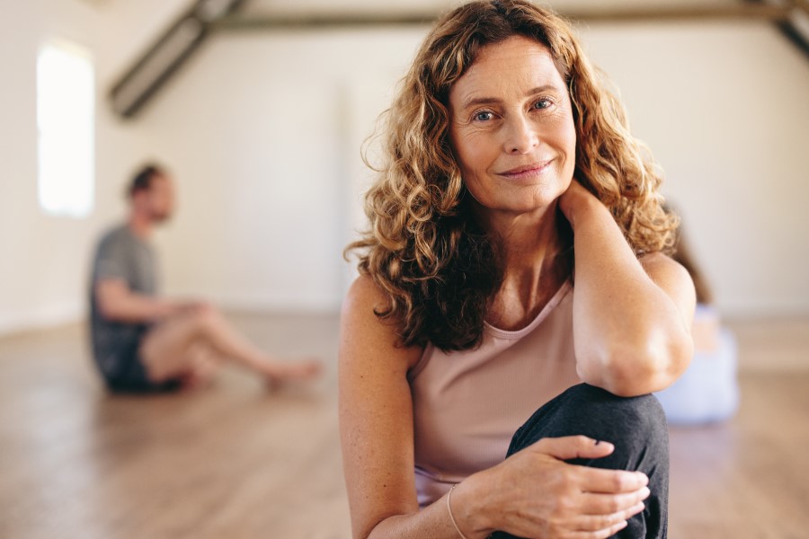 A woman with long hair smiles while sitting on the floor of a yoga, one knee to her chest, one hand behind her neck.