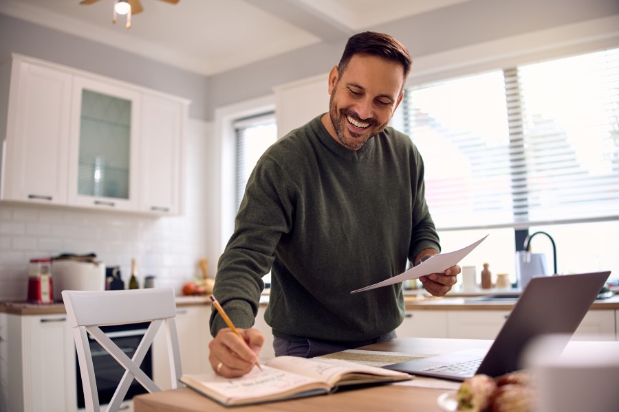 Man at kitchen table writing goals in a book with laptop open