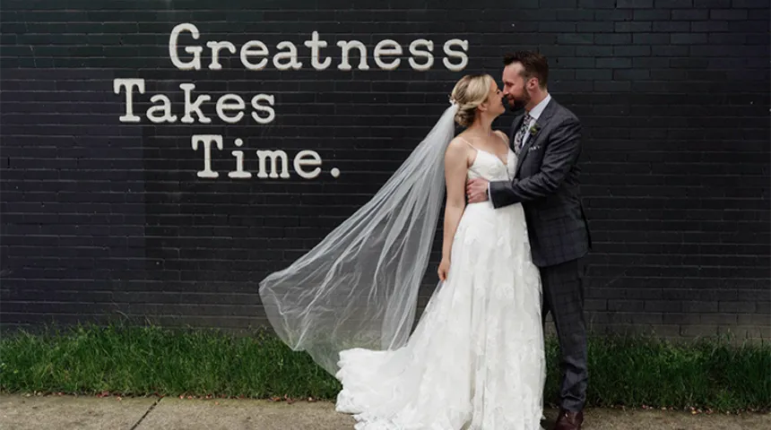 Husband and wife pose in an embrace for an outdoor wedding photo in front of a black, brick wall with Greatness Takes Time painted on it.