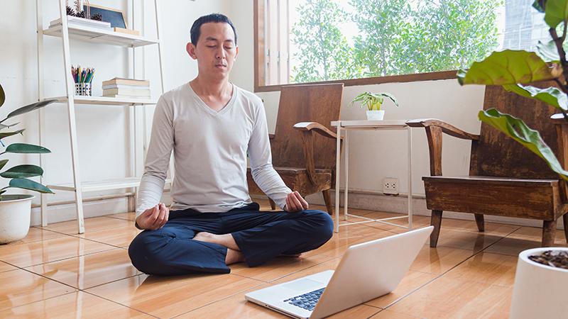 A man sits cross-legged on the floor, eyes closed, back straight.  He has a laptop in front of him.
