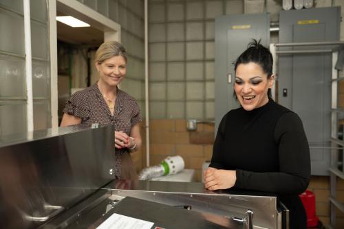 Two women stand in a hospital kitchen. Both look down on a piece of equipment, smiling.