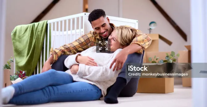 An expecting couple sits on the floor of a a room, with a crib, boxes and houseplants behind them.