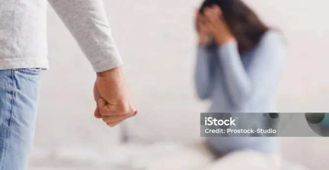 A man's clenched fist is across the room from a scared pregnant woman sitting on the foot of a bed.