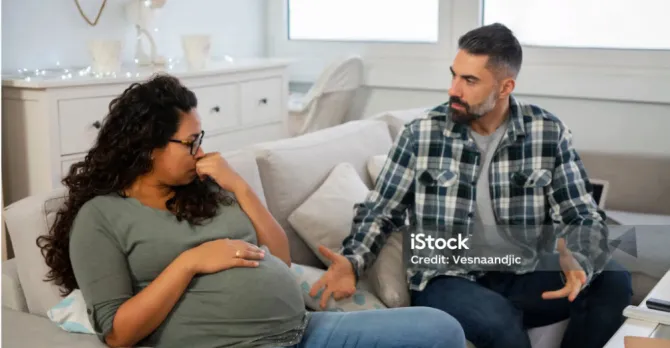 Expecting parents on a sofa.  The pregnant woman is not pointing toward the man, but she looks to the side at him and she looks sad.