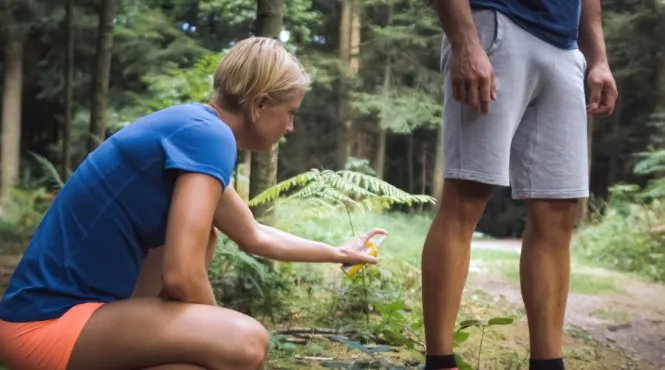 Woman bending down putting bug spray on a man's legs while hiking in the woods.