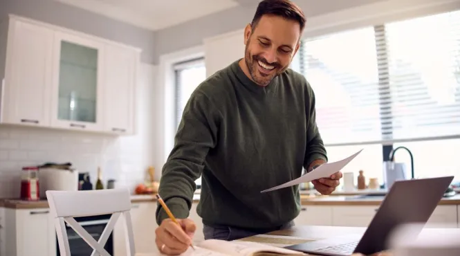 Man at kitchen table writing goals in a book with laptop open