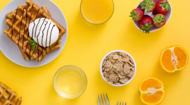 Waffles, cereal, fruit, and juice are shown from above on a yellow table.