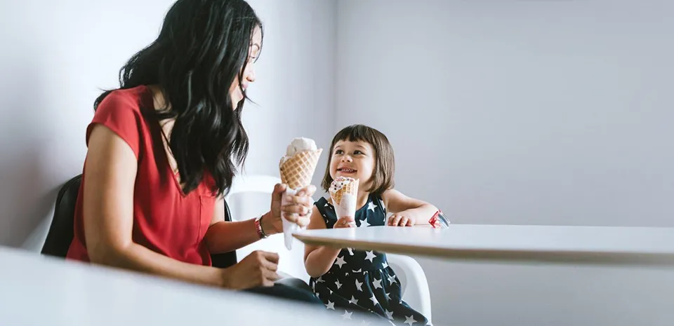 A woman and her daughter sit at a table, eating ice cream cones and having a good conversation.