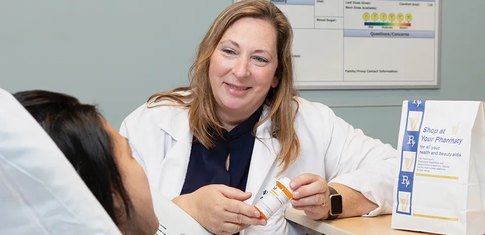 A pharmacist delivers a bag with prescription medication and speaks to the patient who is lying in bed.