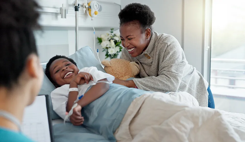 A parent smiling and sitting with their child, who is smiling in a hospital bed.