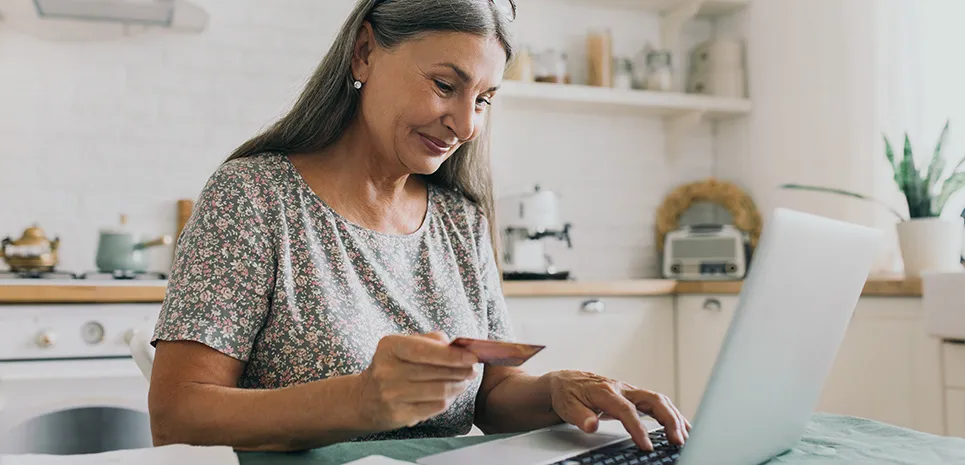 A woman sits at her kitchen table and uses her credit card to pay a bill on her laptop.
