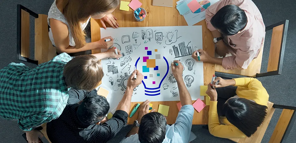 A top-down view of a group of people sitting around a table. The group is writing ideas on a big piece of paper on the table.