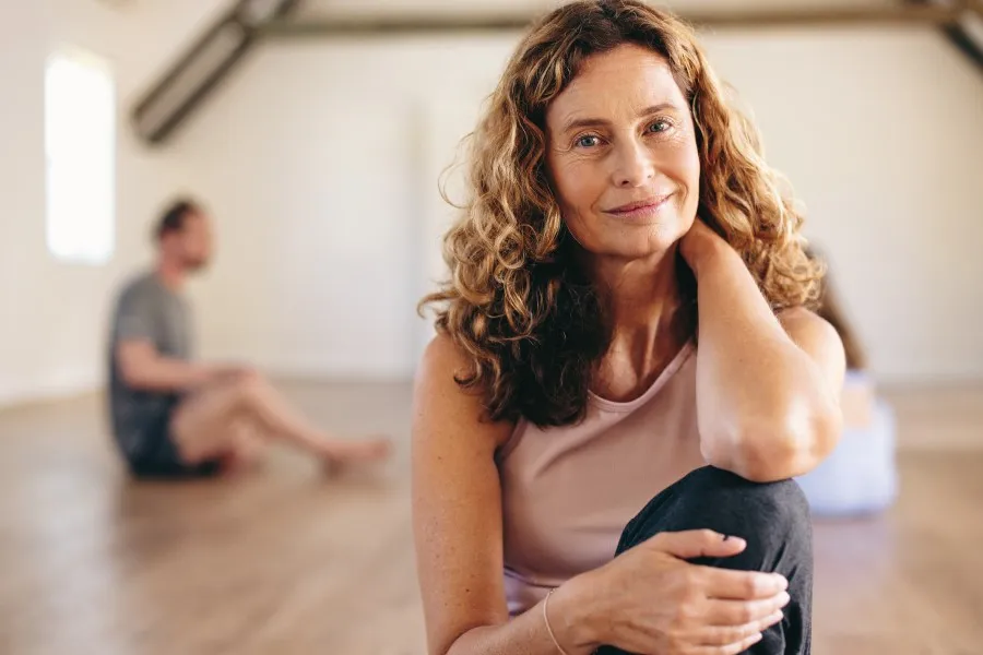 A woman with long hair smiles while sitting on the floor of a yoga, one knee to her chest, one hand behind her neck.
