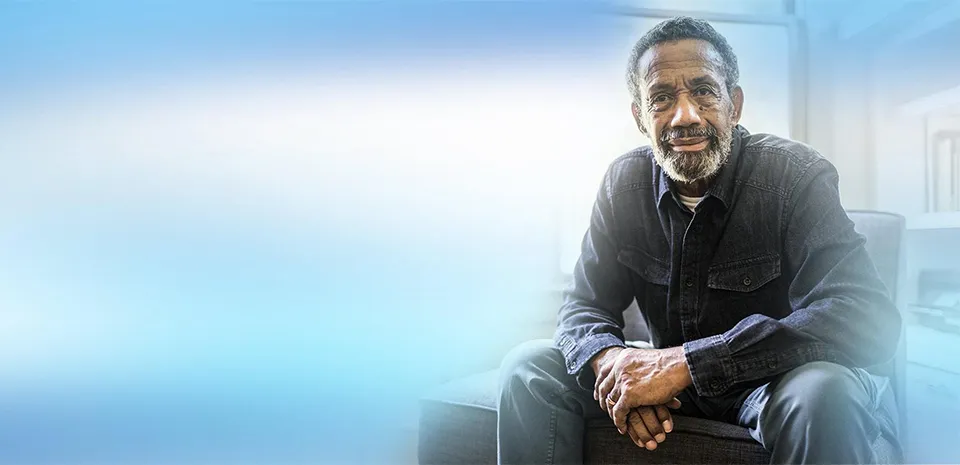 A senior man sits in a living room, looking toward the camera.