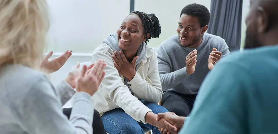 People sitting in a circle are acknowledging an accomplishment for a woman in the group by clapping while someone shakes her hand.
