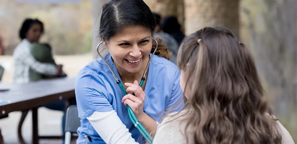 A health care professional examines a pediatric patient in an park using a stethoscope.