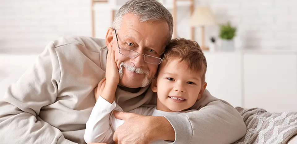 An older man sits on a couch with a young child. The man has his arm around the child while the two both look at the camera.