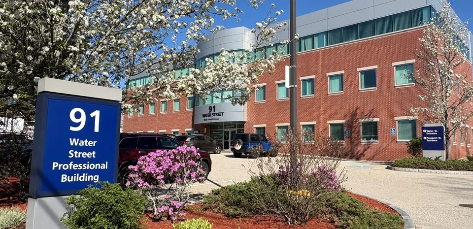 The exterior of the brick building at 91 Water Street in Milford, Mass. on a sunny day. Flowers are blooming on trees.