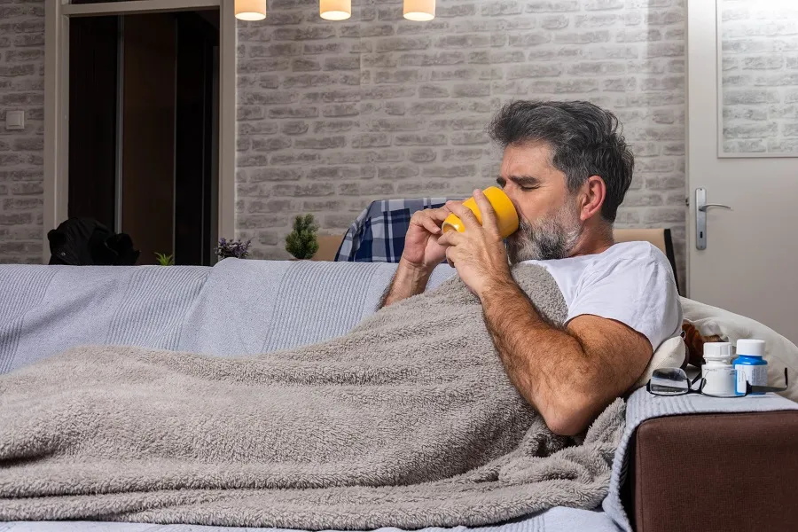 A man is under a blanked on a couch and drinking from a mug.