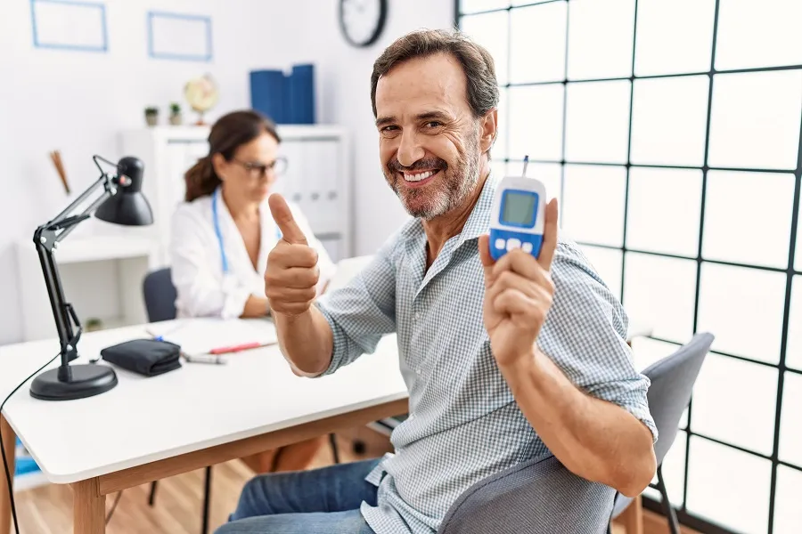 A smiling man is holding a blood glucose meter and giving a thumbs up while in a doctor's office.