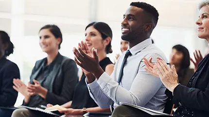 A group of people are seated in a classroom and are clapping while facing the front of the room.