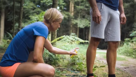 Woman bending down putting bug spray on a man's legs while hiking in the woods.