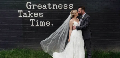 Husband and wife pose in an embrace for an outdoor wedding photo in front of a black, brick wall with Greatness Takes Time painted on it.