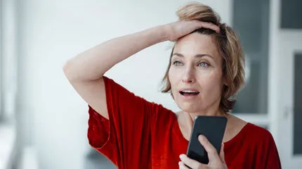 Woman on mobile phone is pushing her hair back off her forehead and looking dismayed.