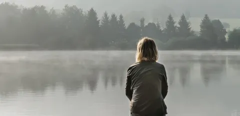 A woman sits on a dock, looking across the water through a thick fog.