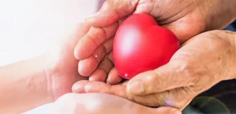 A senior's hands hold a red, heart-shaped object, while a young person's are below, holding the senior's hands.