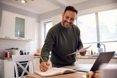 Man at kitchen table writing goals in a book with laptop open