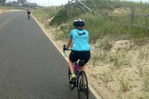 A woman in a blue shirt is riding a bike along a sandy road.