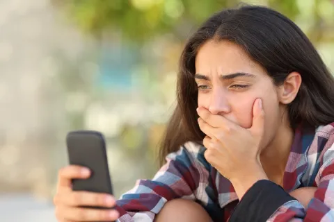 A teen is covering their mouth while looking at a cell phone.