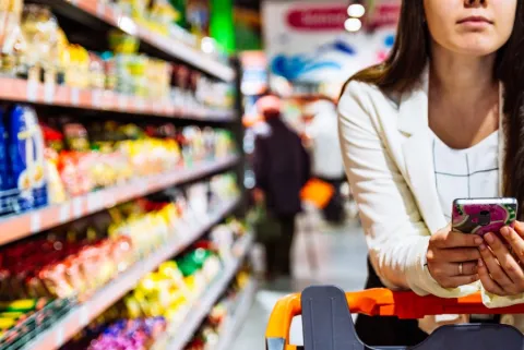 A closeup shows a woman on her cell phone shopping in a grocery store.
