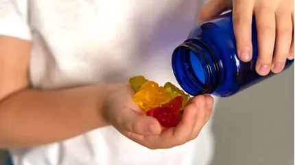 A child holds gummies in one hand and a blue screw-top bottle in the other.