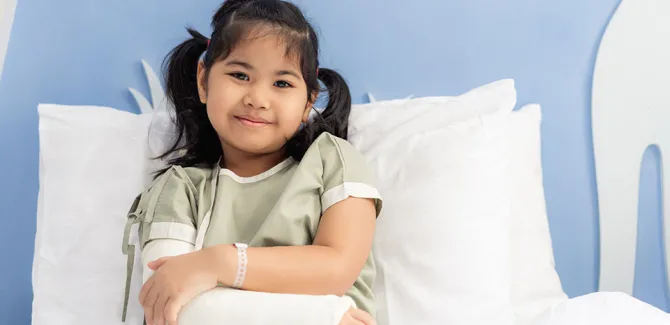A child in a hospital bed wearing a cast on their right arm, looking at the camera.