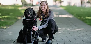 Valentina, black Labrador Retriever on the left, poses with a caregiver outside in the shaded walkway of an outdoor space.