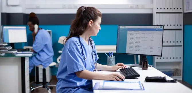 A nurse sitting at a computer station, looking to a computer at the right. In the background another nurse is looking at a computer on their left.