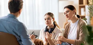 A mother and daughter sit together on a sofa, facing a man on a chair who is taking notes.
