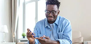 A young black man sits on a chair in his living room doing a finger stick test for diabetes.