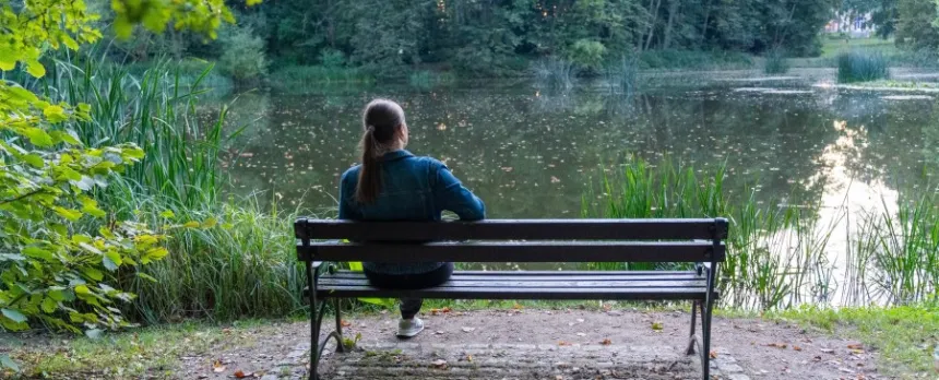 woman sitting on bench looking at trees and pond practicing mindfulness in nature