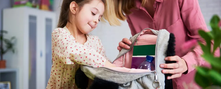 A young girl puts things into her backpack while her mother holds it open on a countertop.