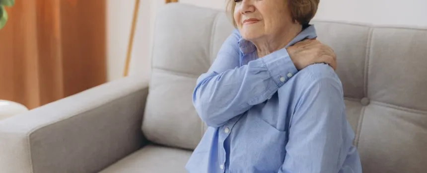 Woman sitting on a couch holding left shoulder wondering if her pain could mean a heart attack