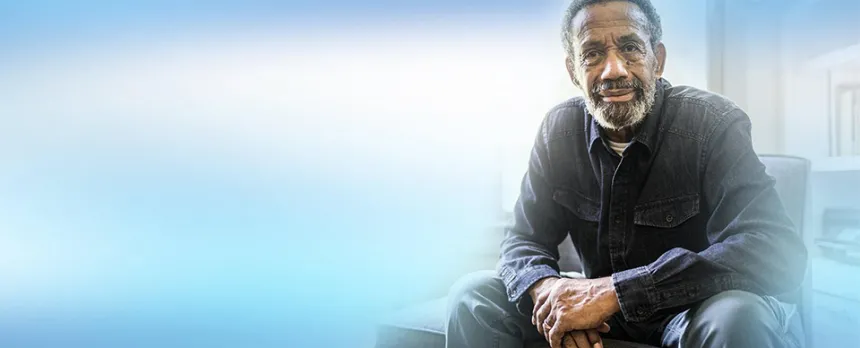 A senior man sits in a living room, looking toward the camera.