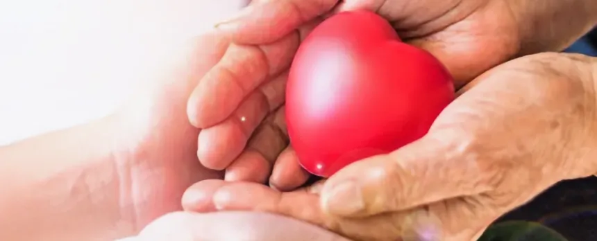 A senior's hands hold a red, heart-shaped object, while a young person's are below, holding the senior's hands.